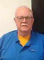 A man named Bob Linde wearing a blue shirt and glasses, standing in front of a black wall.