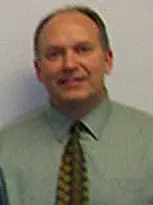 A man named David, wearing a green shirt and tie, stands confidently in front of a white wall.