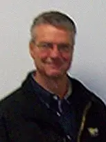 A man named Kevin, wearing a black jacket and glasses, stands in front of a white wall.