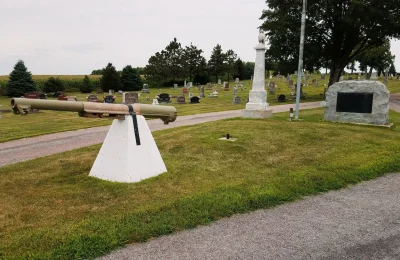 A cannon rests on a grassy field beside a cemetery, evoking a sense of history and remembrance.